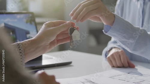 Hands of female real estate agent giving keys to client over office desk with architectural plans and laptop during property ownership transfer. Close-up view