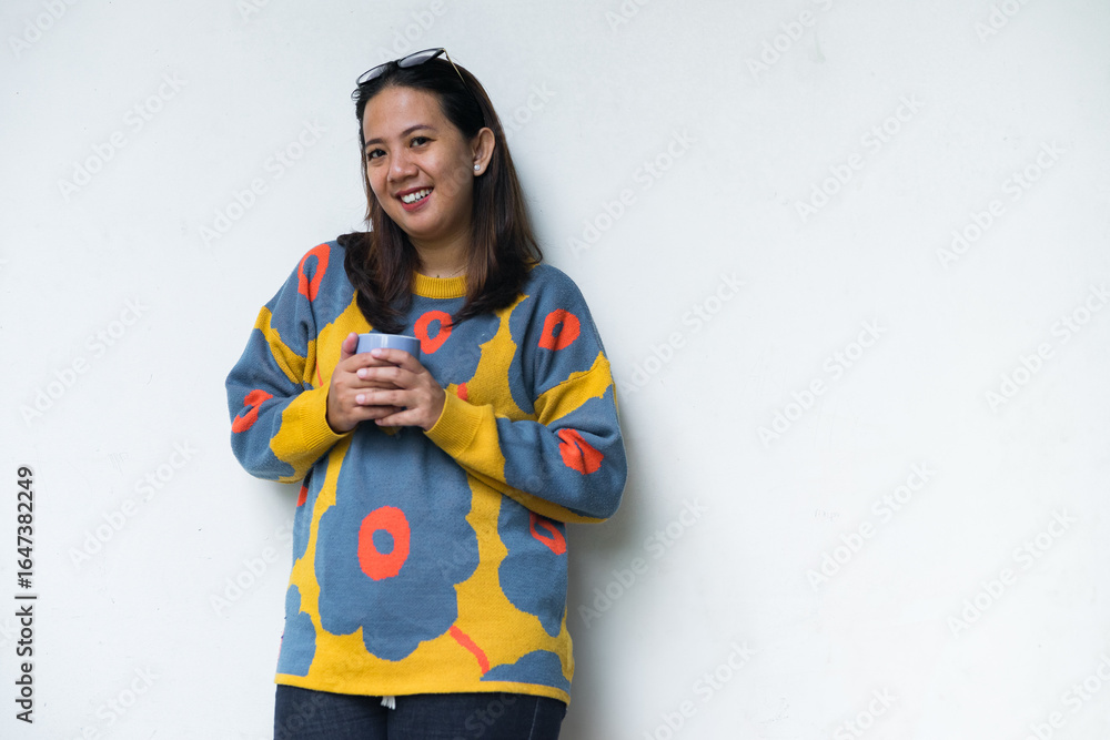 Young Asian woman in a colorful sweater standing by a bright window, holding a coffee mug and smiling, cozy home lifestyle scene. Young Asian woman in a colorful sweater standing by a bright window, holding a coffee mug and smiling, cozy home lifestyle scene.