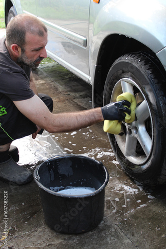 A middle-aged man in black summer clothes and black gloves washes the wheel of a silver car with a soapy sponge.