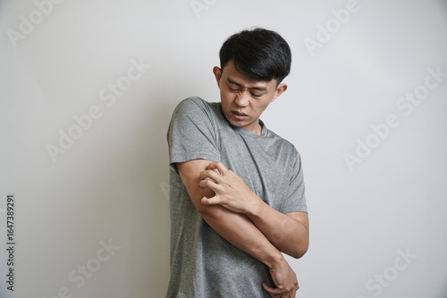 A young Asian man scratches his arm with a grimacing expression, indicating irritation or a skin problem.