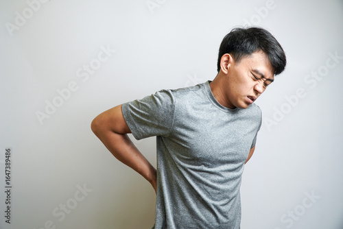 Photography A young Asian man holding his lower back with both hands, with a grimacing expression of pain and discomfort