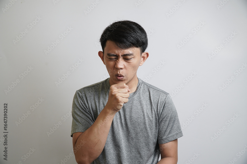 A young Asian man is coughing with his hand covering his mouth, showing symptoms of a cold or flu. A young Asian man is coughing with his hand covering his mouth, showing symptoms of a cold or flu.