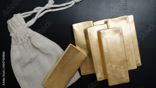 Gold bars arranged on dark surface next to white cloth bag, creating sense of wealth and luxury. shiny metal reflects light, emphasizing its value and allure