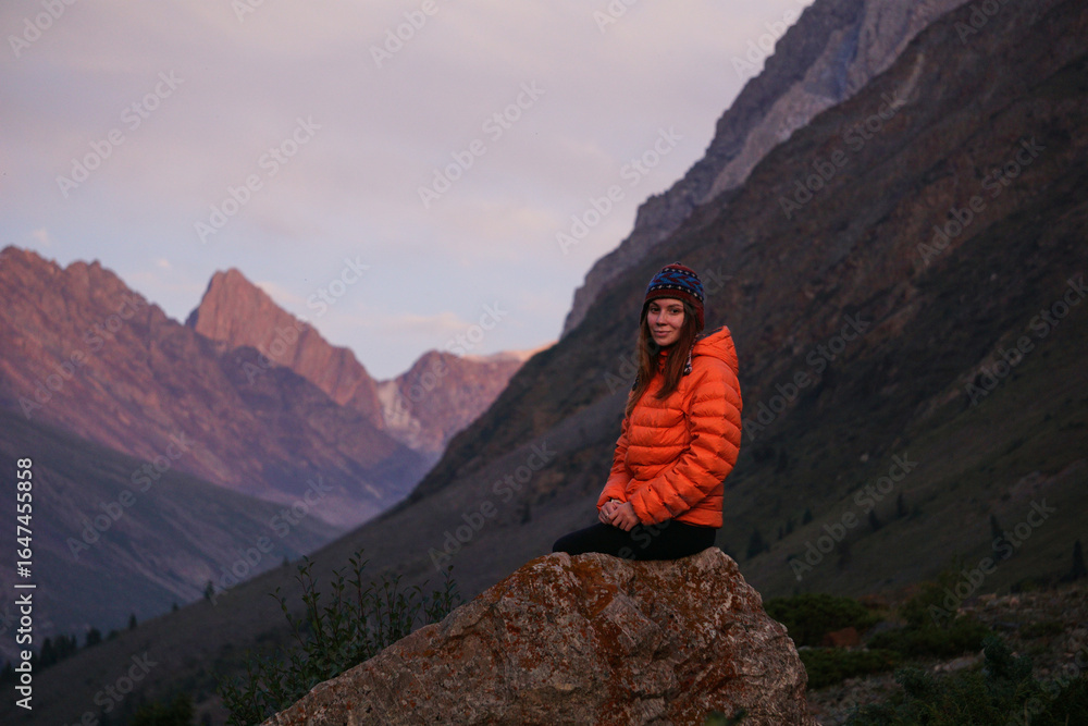 Obraz premium A young woman is sitting on a large rock and enjoying the view of the mountains. Hiker in the mountains during sunset, outdoor activities