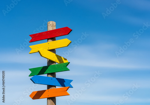 A wooden signpost with multiple staggered, unlabelled, colorful arrows points in various directions under a clear blue sky.