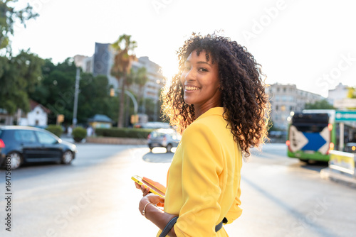 Schilderij op canvas African-American woman in yellow blazer holding smartphone and smiling while walking in city at sunset