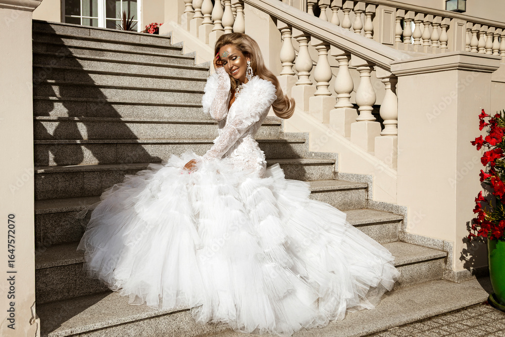 Obraz premium Stunning bride in luxurious white wedding gown with dramatic tulle train posing gracefully on elegant stone staircase in sunlight