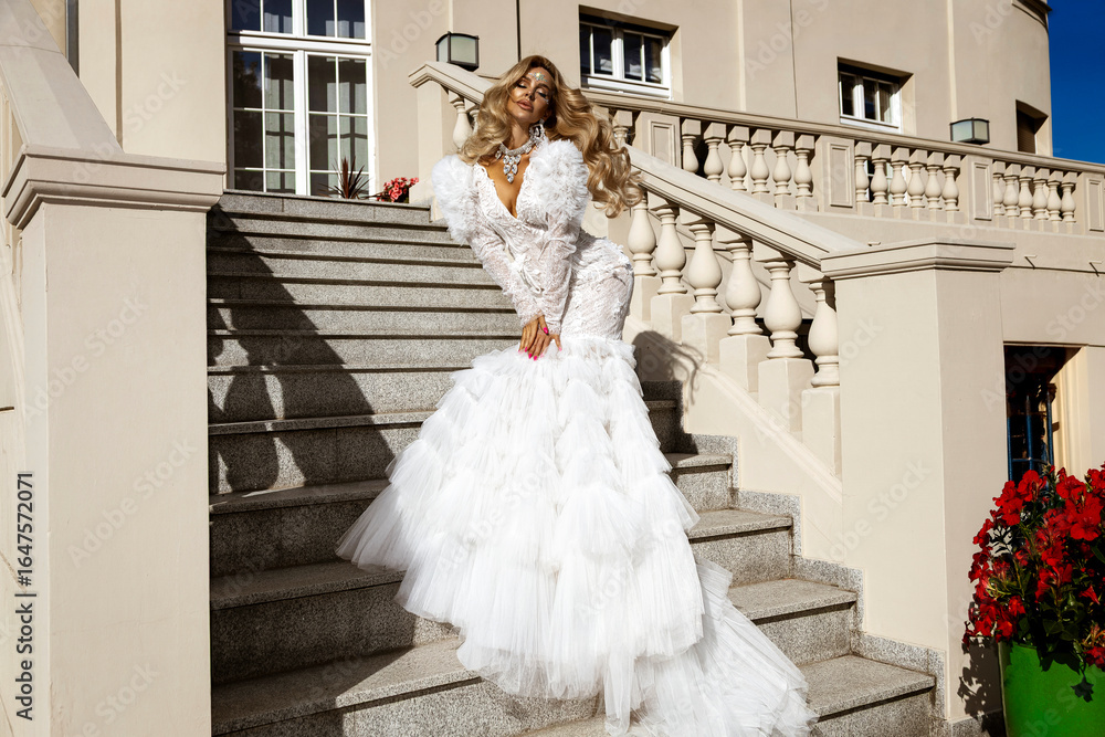 Fototapeta premium Stunning bride in luxurious white wedding gown with dramatic tulle train posing gracefully on elegant stone staircase in sunlight