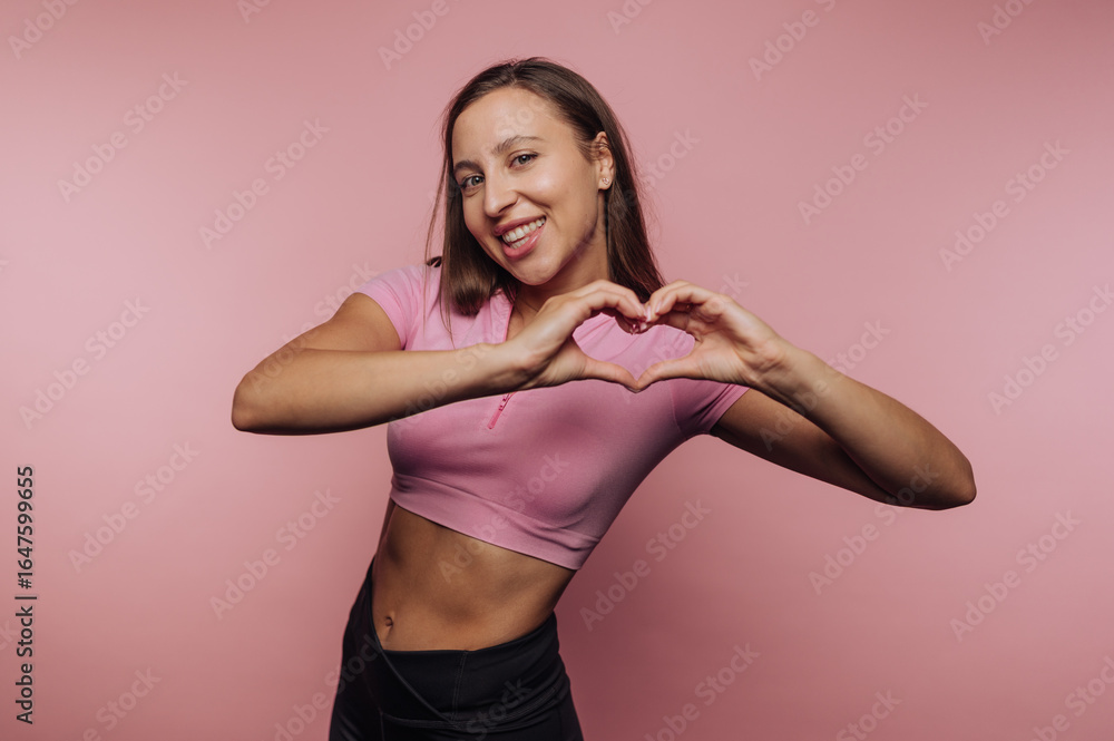 Obraz premium Young woman makes heart shape with hands against pink background while smiling and posing happily in casual outfit