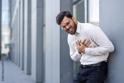 Φωτογραφία A man wearing glasses and a white shirt leans against a wall while holding his chest in pain