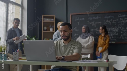 Wallpaper Mural Waist up of Biracial male student working on laptop at classroom desk while teacher and classmates interacting near chalkboard with grammar sentences in background Torontodigital.ca