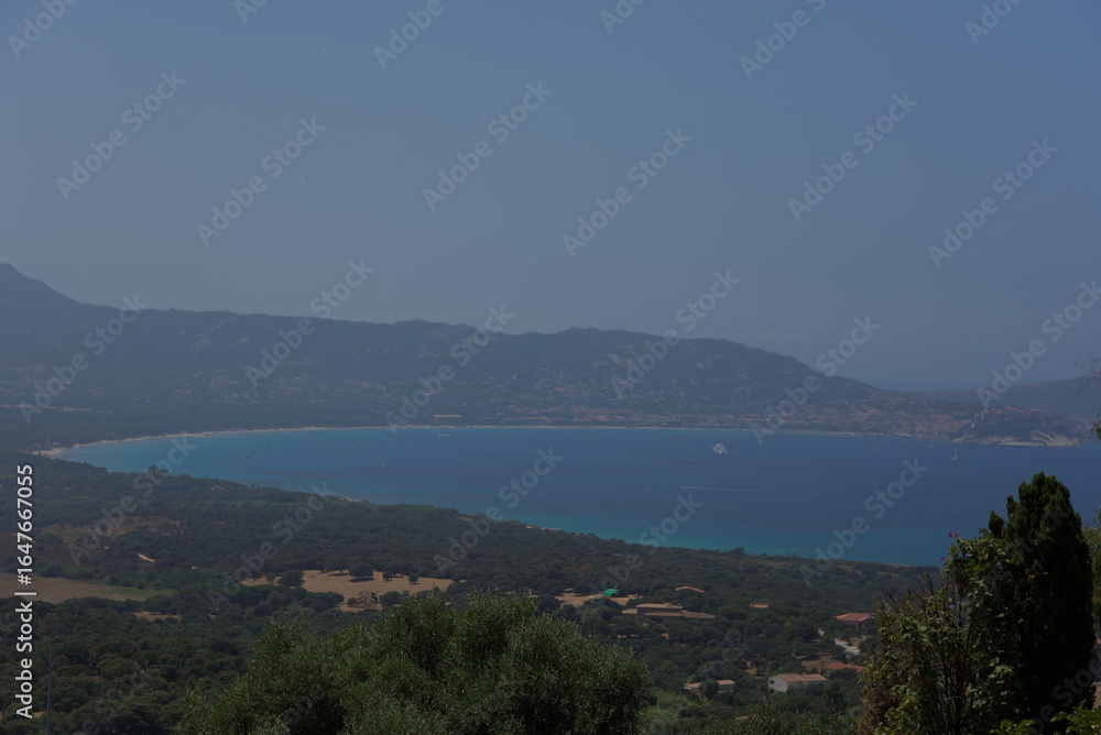 An aerial view shows a calm turquoise bay nestled between lush green hills and a distant mountain range, creating a tranquil and expansive mood. An aerial view shows a calm turquoise bay nestled between lush green hills and a distant mountain range, creating a tranquil and expansive mood.