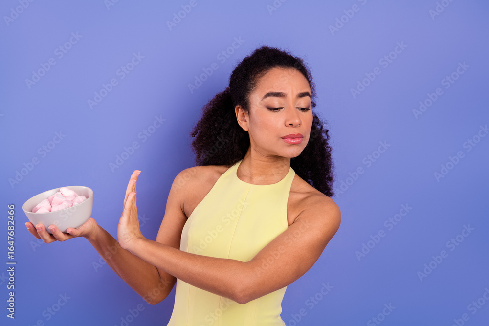 Young woman refuses a bowl of sweets with a serious expression against a purple background, advocating healthy lifestyle Young woman refuses a bowl of sweets with a serious expression against a purple background, advocating healthy lifestyle