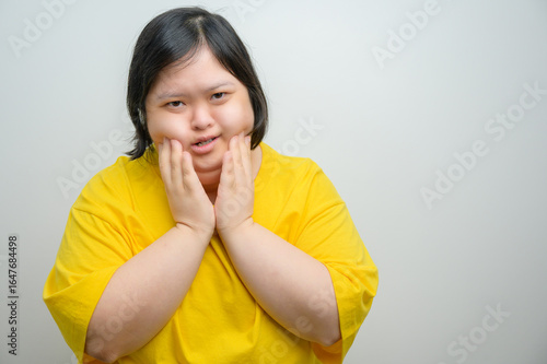 Asian woman with Down syndrome wearing yellow shirt, holding cheeks with painful expression, toothache concept on white background, studio portrait for dental health awareness.