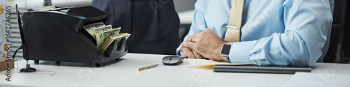 Header of male clerk sitting at desk counting cash with money counting machine in bank office, hands folded on table, wearing business attire, partial body visible