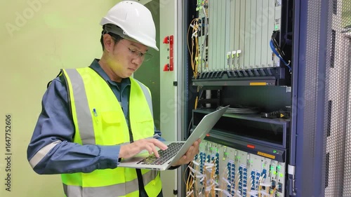 Network technician performing detailed inspection on server rack using laptop in data center with safety helmet and reflective vest ensuring data center safety and efficient network management