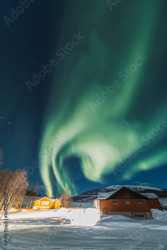 Northern lights light up the entire sky under small wooden cottages with fire inside. Cozy and fantasy evening in Northern Norway