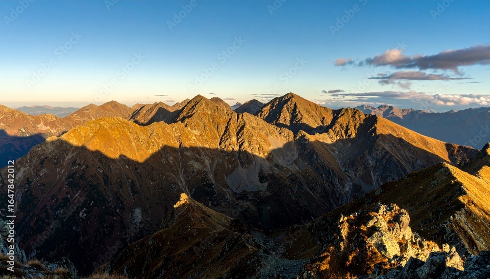 Naklejka premium Panoramic view of rugged mountain peaks in autumn, golden sunlight casting warm glow over rocky ridges, vast landscape with dramatic shadows