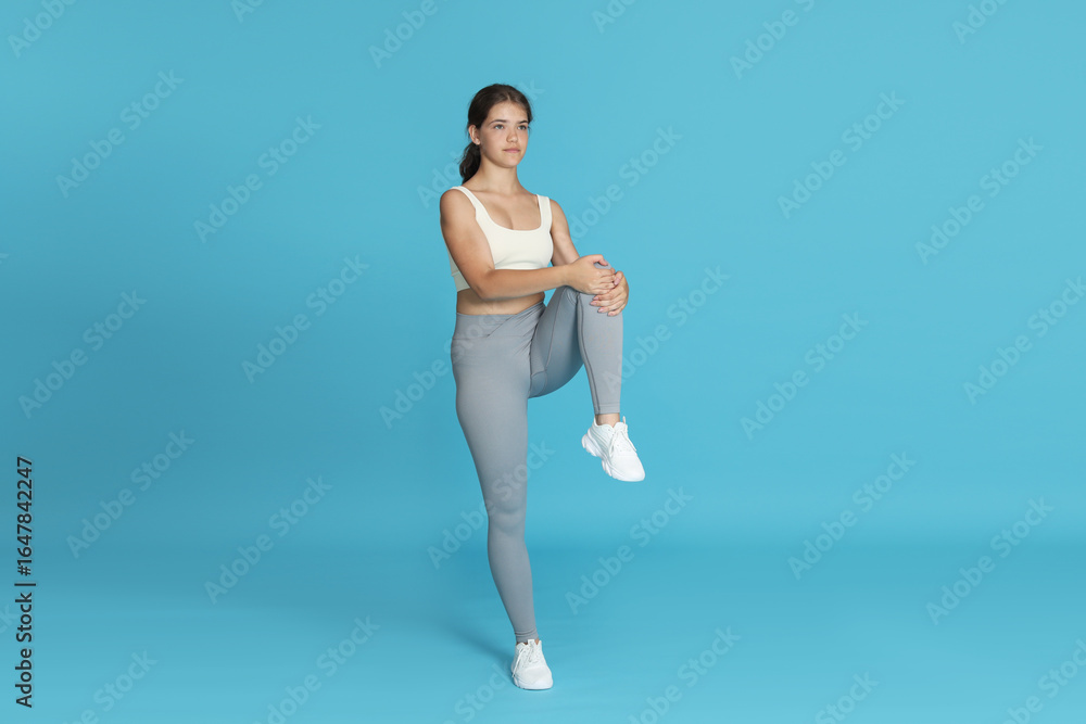 Fototapeta premium Teenage girl in sportswear stretching on light blue background