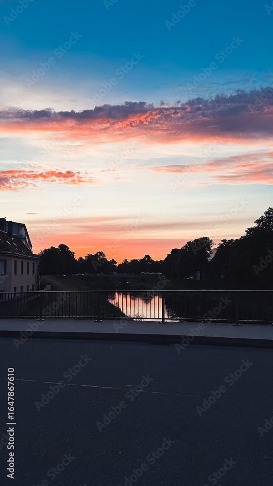 Obraz premium Serene sunset over calm river with vibrant colors reflecting on water, framed by trees and buildings, creating a tranquil evening atmosphere