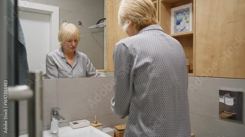 Mature woman performs her daily skincare routine in the bathroom. She applies a facial toner or lotion with a cotton pad for cleansing, promoting anti-aging effects and a fresh healthy skin.