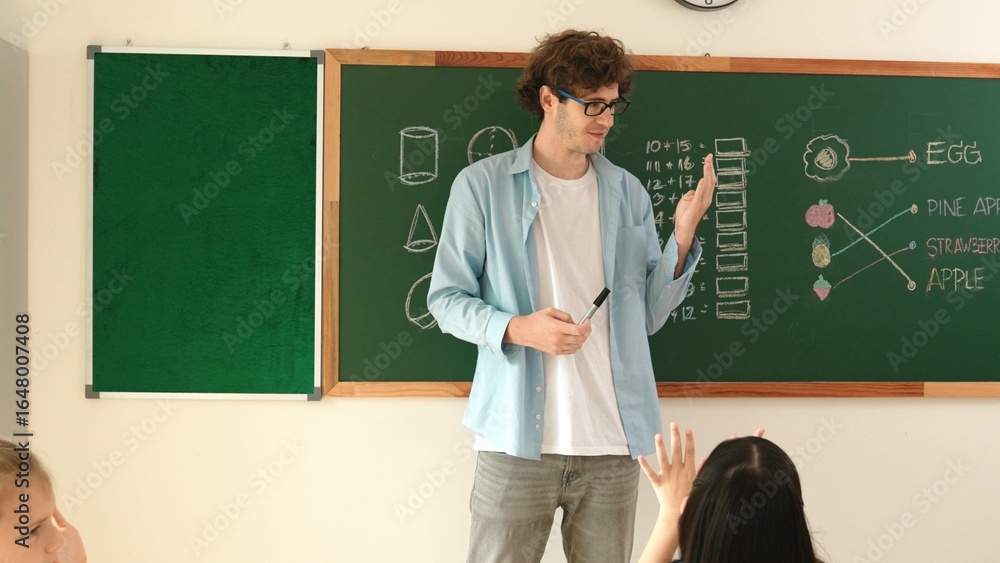 Professional mentor looking around classroom while standing in front of blackboard with math formula or theory. Diverse elementary student learning or studying and sitting in Math lesson. Pedagogy. Professional mentor looking around classroom while standing in front of blackboard with math formula or theory. Diverse elementary student learning or studying and sitting in Math lesson. Pedagogy.