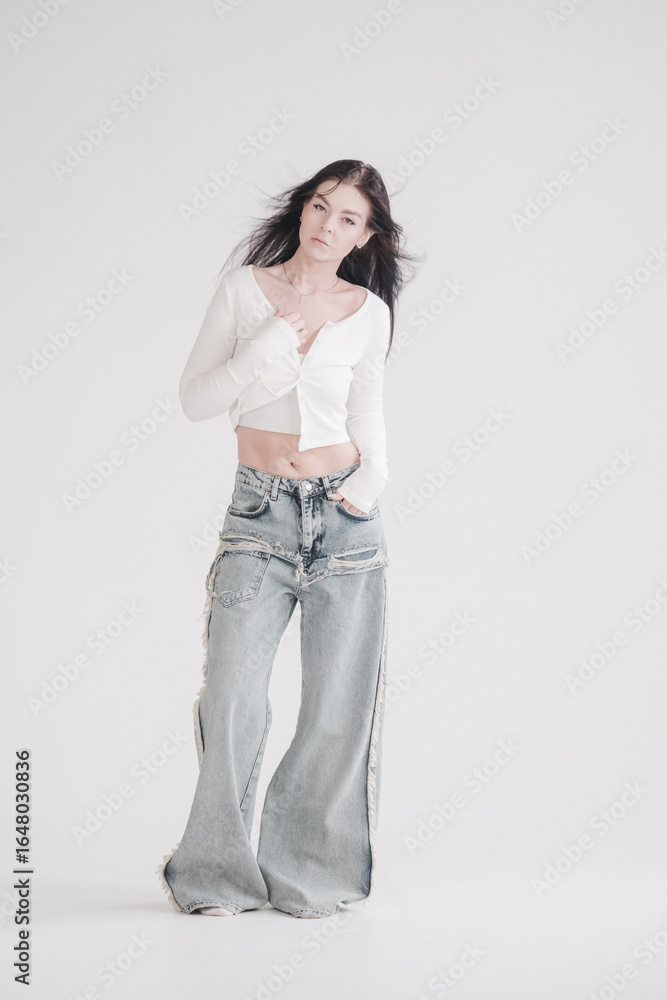 Pretty young fashion sensual woman posing on white wall background dressed in white top and jeans style outfit. Stylish fashionable girl in basic clothes. Her hair is blowing in the wind. Mockup Pretty young fashion sensual woman posing on white wall background dressed in white top and jeans style outfit. Stylish fashionable girl in basic clothes. Her hair is blowing in the wind. Mockup