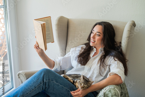Young woman sits in a cozy armchair and reads a book, with a cat in her arms. Autumn evenings at home.