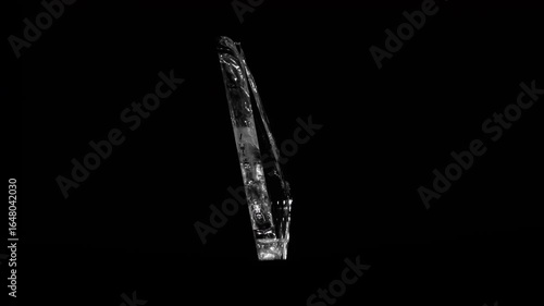 A thin, elongated shard of broken glass rotates gracefully against a black backdrop, catching light in its edges.