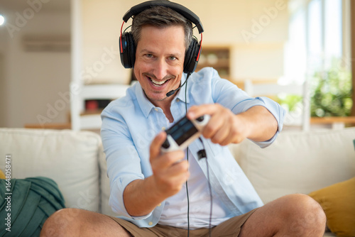 Smiling man wearing headphones and playing video games at home, enjoying online gaming.
