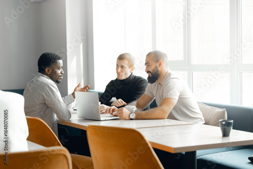 Three diverse men engaged in collaborative meeting in well-lit modern office. focused interaction around laptop reflects teamwork, business strategy, and professional communication.