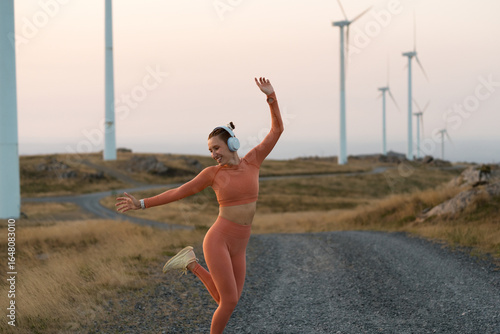 Young sportswoman listening to music and dancing near wind turbine farm