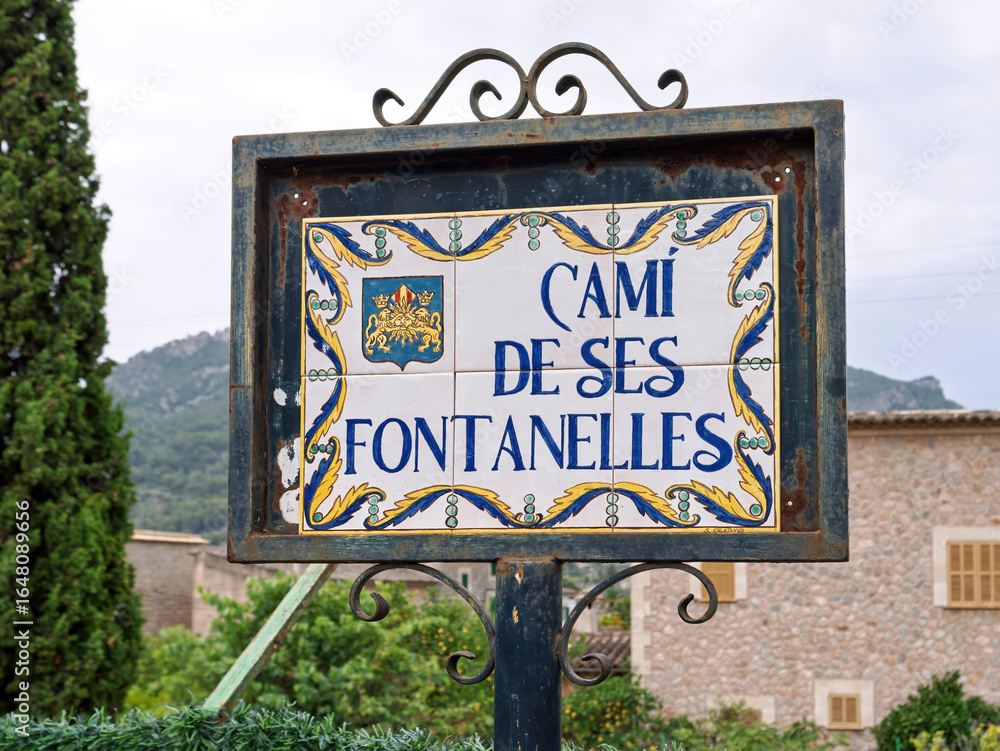 Obraz premium Decorative ceramic street sign for Cami de ses Fontanelles in Soller, Mallorca, framed in wrought iron with scenic mountain backdrop.