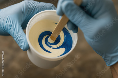 Mixing Colors: A close-up shot revealing the intricate dance of color as blue and white hues are meticulously blended, with a wooden stir stick within a plastic cup. 