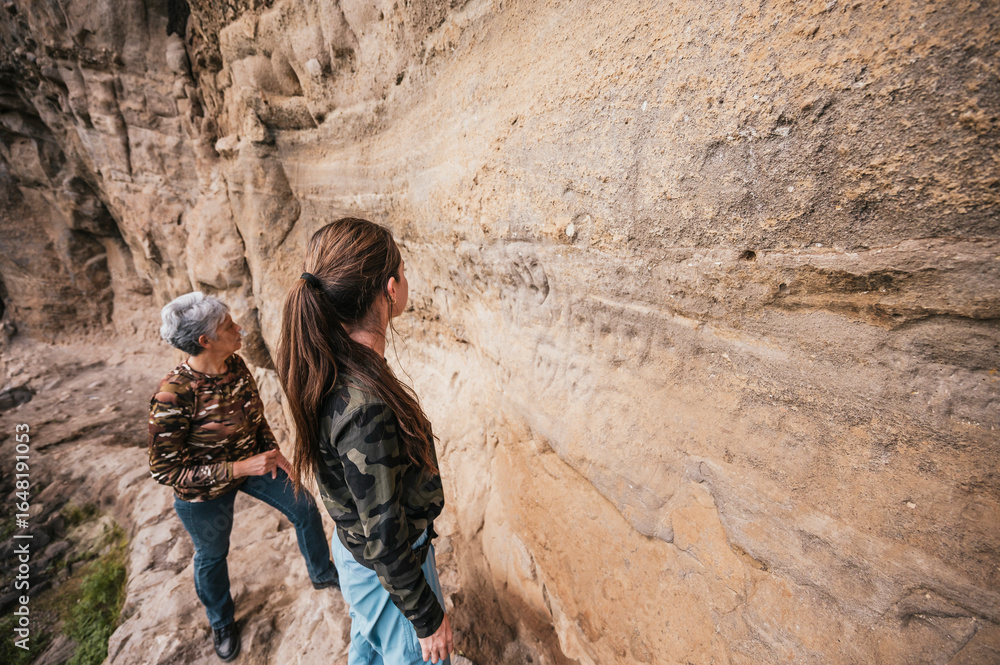 Naklejka premium Mother and daughter dressed in hiking clothes, walking along a cliff observing cave paintings.