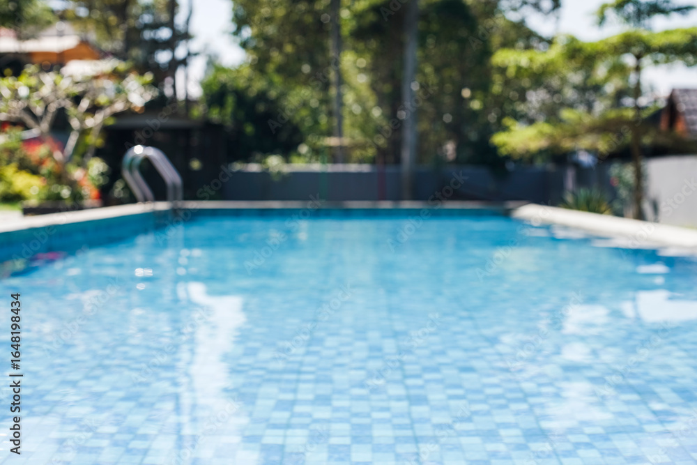 Naklejka premium A beautifully blurred swimming pool with blue tiles and sparkling water, surrounded by lush greenery, ideal for a serene summer escape or luxurious travel.