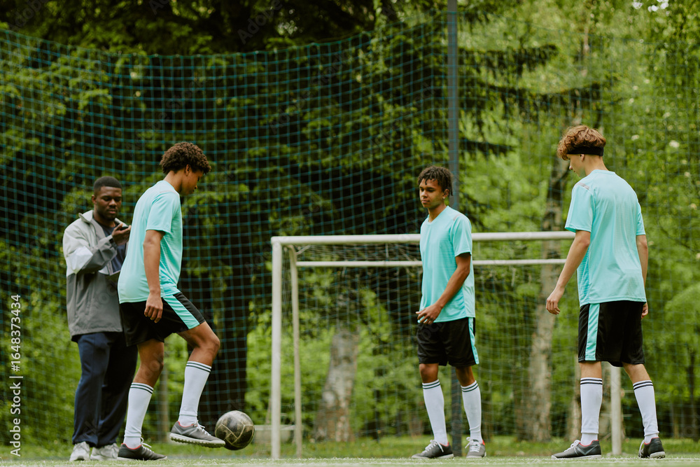 Obraz premium Black male coach instructing three teenage boys, two Caucasian and one Black, practicing soccer on outdoor field, one teenager controlling ball with foot near goalpost, trees in background