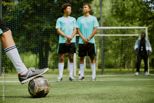 Teenage boy preparing to kick soccer ball during free kick while two teenage boys, one Black and one Caucasian, standing in defensive wall on outdoor soccer field
