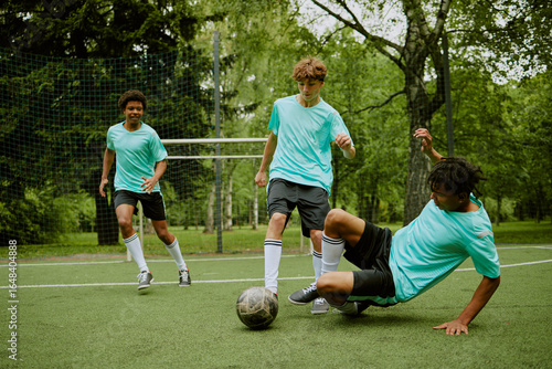 Three teenage boys playing soccer on outdoor field  one Black teenager sliding to tackle ball while others running and focusing on game action