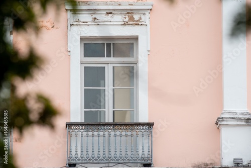 Ornate balcony with a vintage window on a pastel pink building facade, framed by blurred greenery