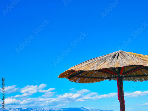 Straw beach umbrella under blue sky with mountains in background
