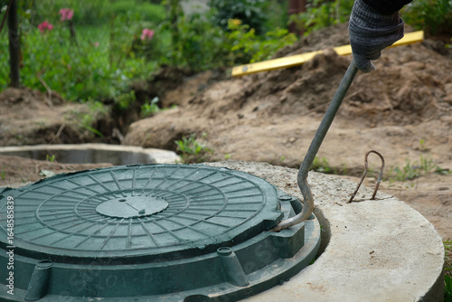 Opening a green septic tank manhole cover with a metal hook in a private house yard for inspection and maintenance of a local sewage system, wastewater management, and underground drainage infrastruct