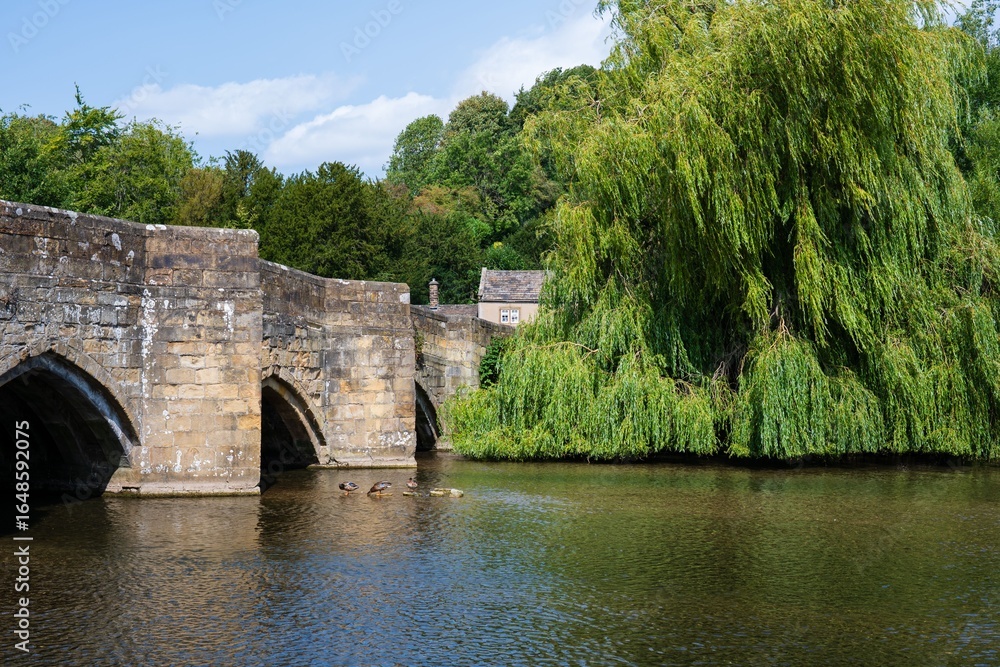 Fototapeta premium Bakewell Derbyshire.Stone bridge over a serene river by lush green trees under blue sky.