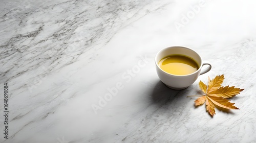 White ceramic cup with golden liquid beverage and autumn leaf on marble surface.