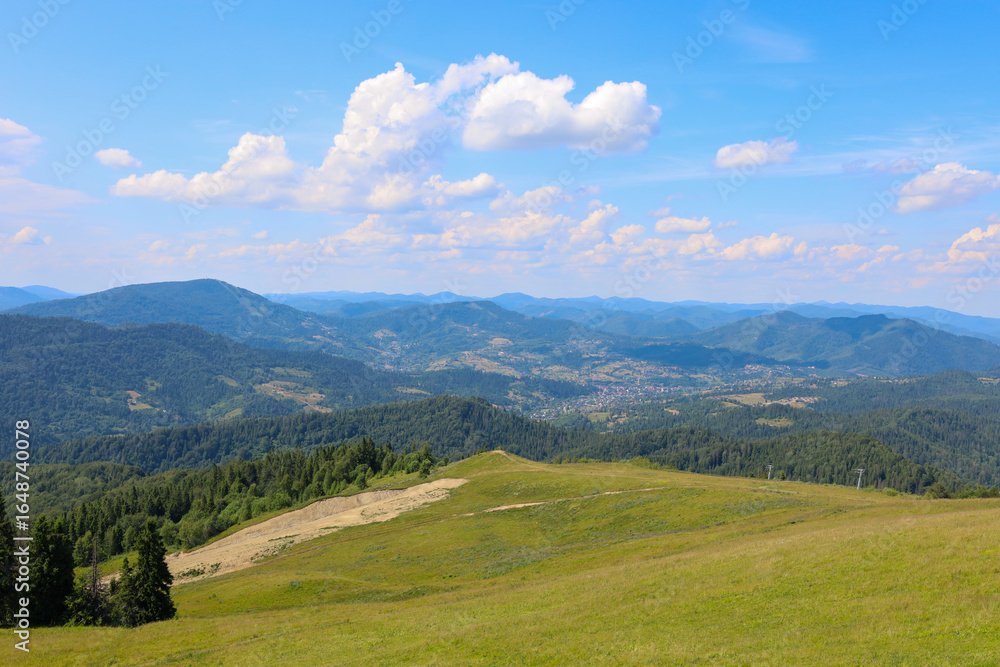 Naklejka premium A summer mountain landscape where the foreground is a green hill, offering a breathtaking panorama of wooded mountain ranges. In the distance, surrounded by greenery, a small village is visible.