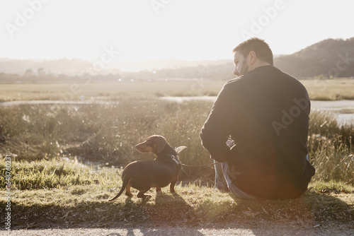 Man in black long sleeved shirt and blue jeans sitting out in nature next to a lake with a brown dachshund in a harness and on leash engaging with his pet dog. Sun flare. Lake and grass