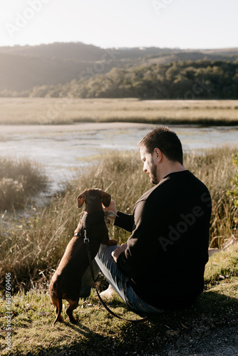 Man in black long sleeved shirt and blue jeans sitting out in nature next to a lake with a brown dachshund in a harness and on leash engaging with his pet dog. Sun flare. Lake and grass