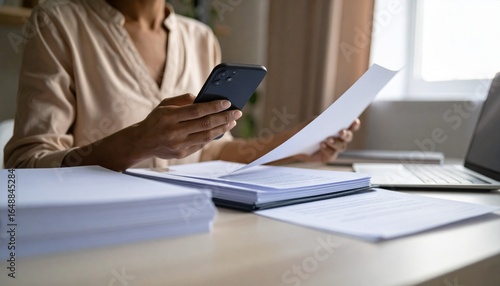 A person flipping through documents on a desk and checking their smartphone