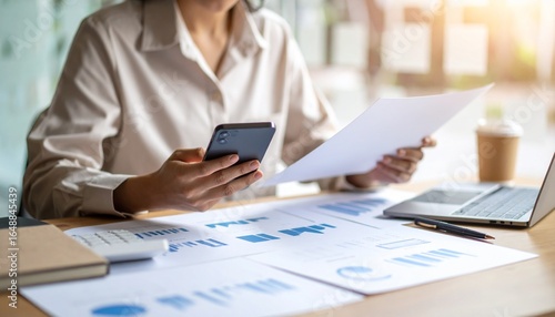 A person flipping through documents on a desk and checking their smartphone