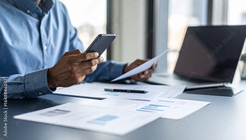 Fototapeta premium A person flipping through documents on a desk and checking their smartphone